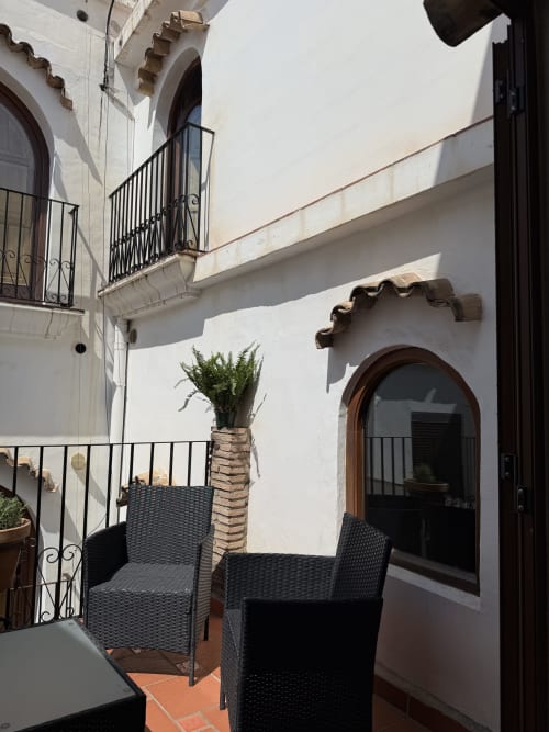 Acogedora terraza de Hotelito Oliva con sillas de mimbre negro, palmera en maceta y puerta arqueada con vistas a la ciudad de Oliva