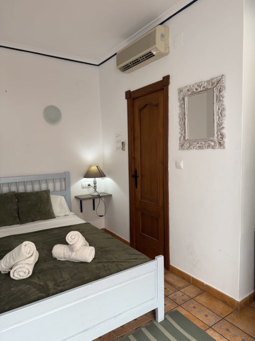 Acogedora habitación del Hotelito Oliva con cama blanca, ropa de cama verde, puerta de madera, aire acondicionado y lámpara de noche en Oliva,