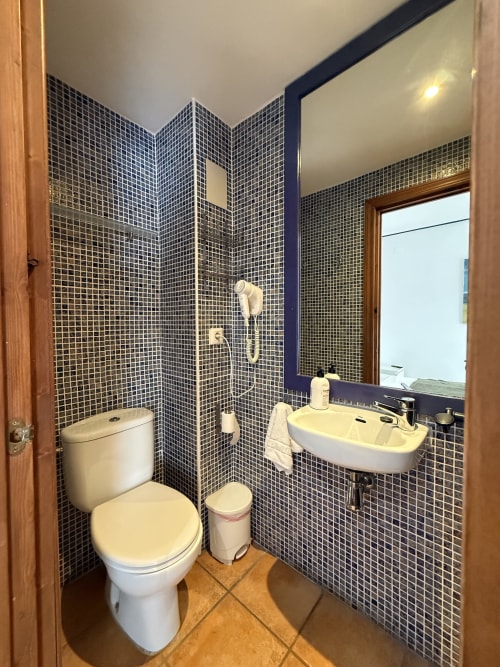Baño moderno con azulejos negros en mosaico, lavamanos blanco, inodoro y ducha en Hotelito Oliva Pensión de Oliva