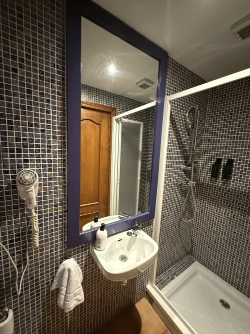 Baño moderno del Hotelito Oliva Pensión con paredes de azulejos oscuros, lavabo blanco, espejo y cabina de ducha