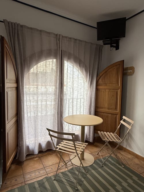 Habitación luminosa en Hotelito Oliva Pensión con mesa redonda, dos sillas, ventanas arqueadas con cortinas translúcidas, puertas de madera
