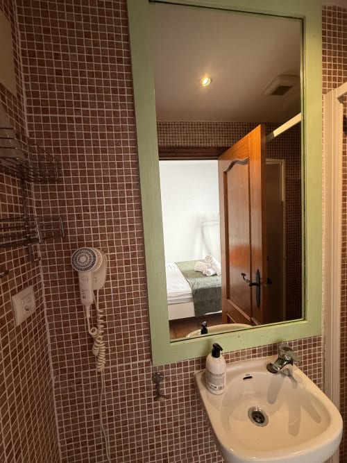 Baño del hotel Hotelito Oliva con paredes de azulejos, lavamanos, espejo y secador de cabello, vista del dormitorio reflejada en el espejo