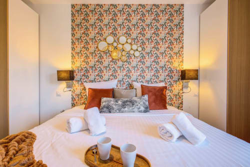 Luxusschlafzimmer in Cannes mit gemusterter Tapete, orangefarbenen Samtkissen, goldenem Spiegel und Willkommensmitnahmen auf dem Bett