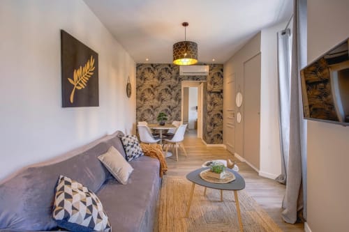 Elegantes Wohnzimmer einer Cannes-Wohnung mit grauem Sofa, Blattgoldkunst, gemusterter Tapete und Essbereich durch Bogenöffnung