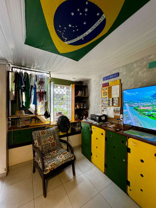 Área comum do Hostel New Ipanema decorada com bandeira brasileira, móveis verde e amarelo, mesa com TV e cabideiro de roupas