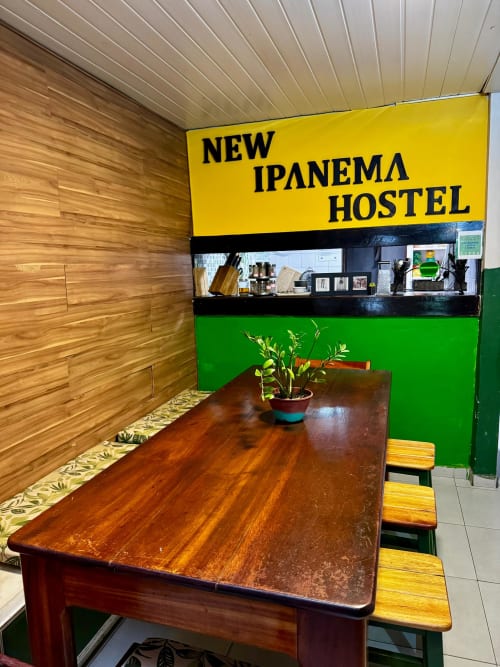 Área comum do New Ipanema Hostel com mesa de madeira, paredes verdes e sinalização amarela vibrante no Rio de Janeiro