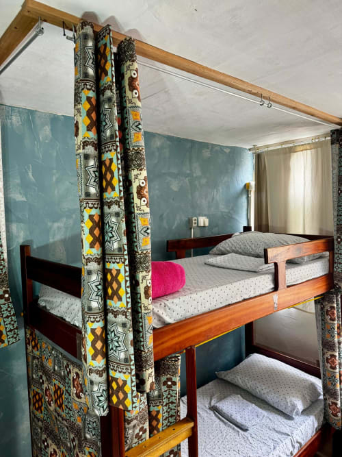 Dormitório feminino do Hostel New Ipanema com beliches de madeira, cortinas estampadas coloridas, paredes verde-azulado e roupas de cama brancas no Rio de