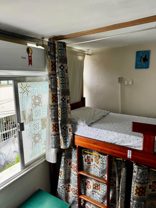Quarto feminino do Hostel New Ipanema no Rio com cama de madeira, cortinas estampadas e vista da janela