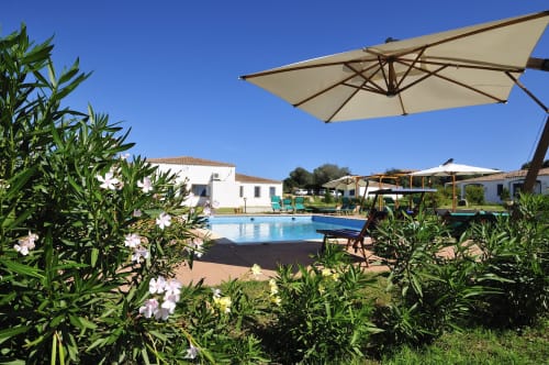 Area piscina del Resort Baia Cea a Bari Sardo con ombrelloni bianchi, lettini, piante fiorite e architettura mediterranea