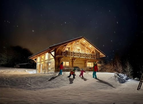 Chalet en bois à Gérardmer sous la neige, familles jouant dehors la nuit, lumières chaleureuses illuminant la scène