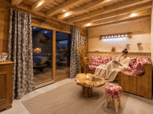 Intérieur cosy d'un chalet alpin avec poutres en bois, canapé fleuri rose, vue sur les montagnes par les portes vitrées du Domaine des Fleurs,