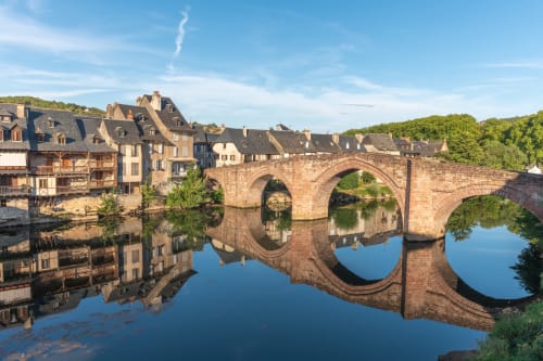 Pont de pierre médiéval aux arches se reflétant dans une rivière calme, bâtiments historiques sur la rive gauche, collines verdoyantes, Estaing