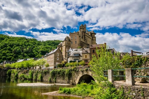 Hôtel château médiéval avec pont de pierre surplombant la rivière à Estaing, France, entouré de verdure luxuriante sous un ciel bleu