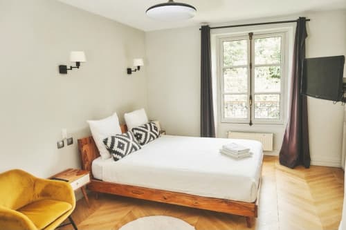 Chambre moderne à la Villa Raspail B&B à Ivry-sur-Seine avec lit en bois, linge blanc, fauteuil jaune et portes donnant sur le jardin