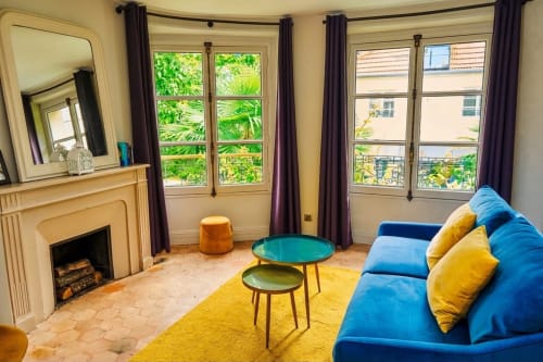 Habitación luminosa con vistas al jardín en Villa Raspail en Ivry-sur-Seine, sofá azul, chimenea, terraza y exuberante vegetación