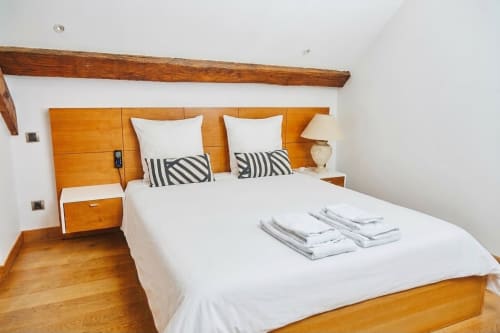 Chambre moderne à la Villa Raspail avec tête de lit en bois, linge blanc, coussins rayés, poutre apparente et parquet