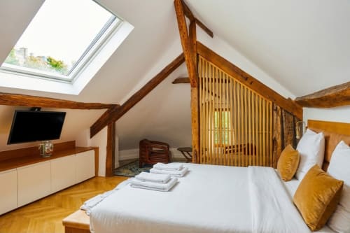 Chambre mansardée élégante à la Villa Raspail avec poutres apparentes, draps blancs, coussins dorés, puits de lumière et TV