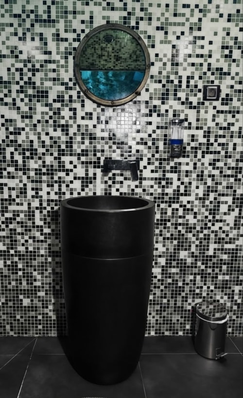 Baño moderno con lavabo cilíndrico negro y portilla circular que muestra agua azul, pared de azulejos mosaico en la Villa