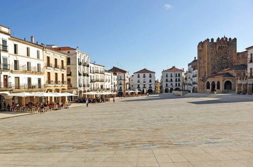Plaza histórica en Cáceres, España con torre medieval, edificios coloniales y mesas de café al aire libre
