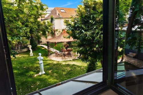 Vista del jardín Villa Raspail desde la habitación Maison Gloriette con exuberante vegetación, glorieta y casa histórica en Ivry-sur-Seine