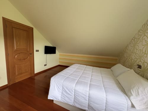Acogedora habitación abuhardillada en Casa Aida con ropa de cama blanca, puerta de madera, TV y papel pintado estampado en Vilaronte, Lugo