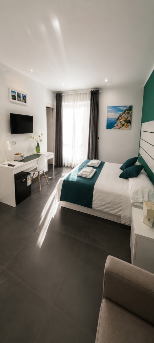 Camera moderna a Soave Rooms a Napoli con letto queen, accenti blu teal, scrivania, TV a schermo piatto e porte vetrate verso il balcone