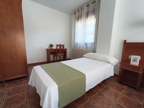 Bright bedroom with wooden furniture, white linens, green throw blanket, and terracotta tiles at Hospedería Puerta a los