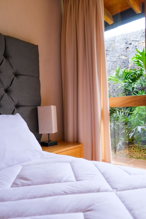 Modern bedroom at La Hacienda Tepoztlán with garden view, white bedding, dark headboard, and terracotta curtains opening to