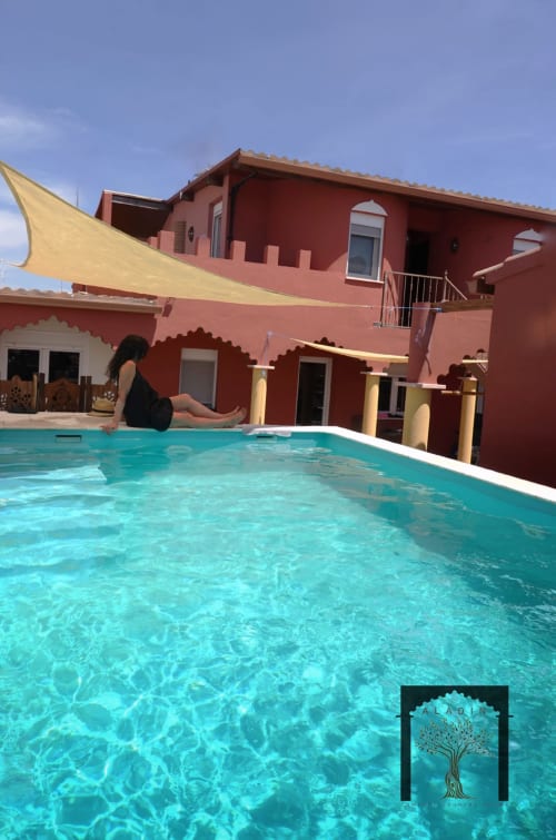 Mulher relaxando à beira da piscina turquesa no Alojamentos Alqueva Monsaraz, edifício vermelho com velas de sombra amarelas e varanda.