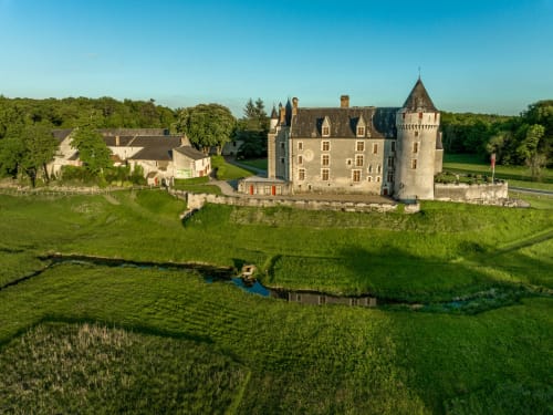 Le Perroquet Bleu castle hotel in French countryside with tower, moat, green grounds and trees under clear sky