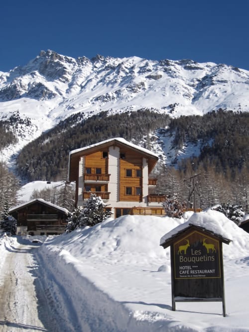 L'hôtel Les Bouquetins en Anniviers enneigé avec montagnes neigeuses et forêts de pins en arrière-plan