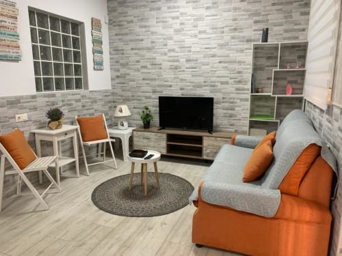 Salón moderno de Milán Soria Luxury en Benidorm con acentos naranjas, sofá gris, TV y paredes de azulejos rayados.