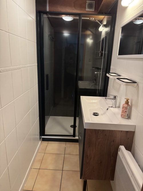 Salle de bain moderne avec douche encadrée noire, vasque blanche et carrelage crème à l'appartement La Guisane