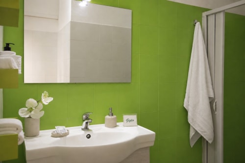 Bright green bathroom with white pedestal sink, mirror, and white tiles at Giglio room in Caltagirone, Sicily