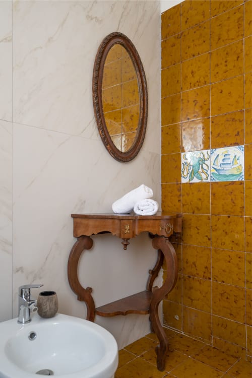 Bagno siciliano con elegante lavabo in legno, specchio ovale, piastrelle Maiolica dorate e asciugamani bianchi presso I colori della Sicilia