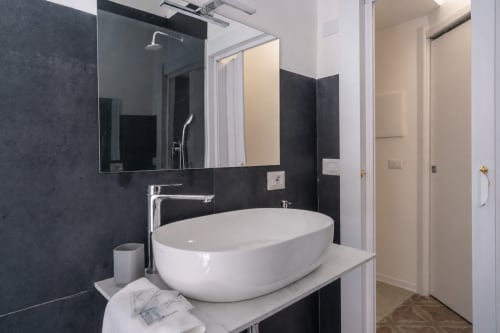 Modern bathroom with oval white sink, black tile walls, mirror, and shower in Ambra room at I colori della Sicilia BnB,
