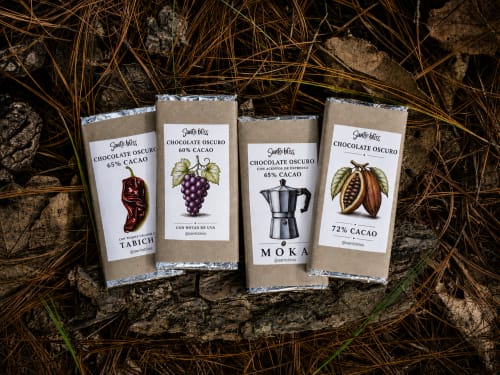 Cuatro paquetes de chocolate negro Santo Bliss exhibidos sobre tierra, con variedades de cacao de Sento SanMa, San Mateo Rio Hondo