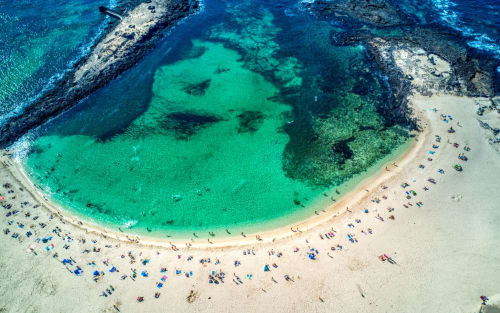 Vista aérea de la playa virgen de El Cotillo con aguas turquesas, arena blanca y sombrillas de colores en la orilla