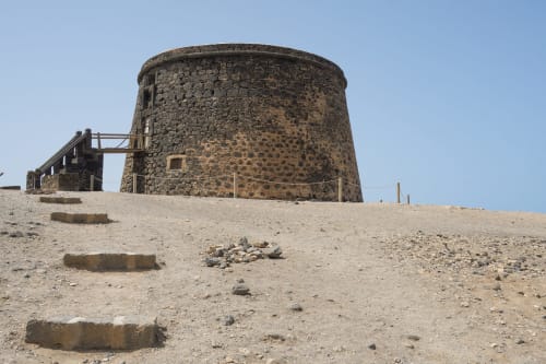 Torre de piedra histórica en El Patrón Casa de Huéspedes, El Cotillo, España con paisaje desértico y estructuras rústicas de madera.