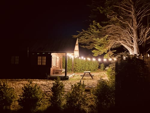 Gîte du Cap Fréhel illuminé la nuit, chalet éclairé, table de pique-nique, guirlandes lumineuses et grands arbres à Plévenon