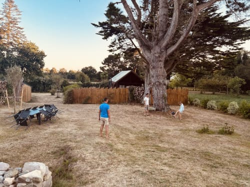 Enfants jouant dans la cour de sable du Gîte du Cap Fréhel avec un ancien cyprès et une cabane en bois à Plévenon