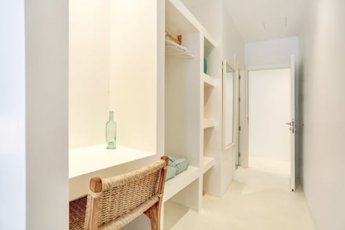 Habitación luminosa y minimalista en El Patrón Casa de Huéspedes con rincón de ventana, silla tejida y estanterías blancas