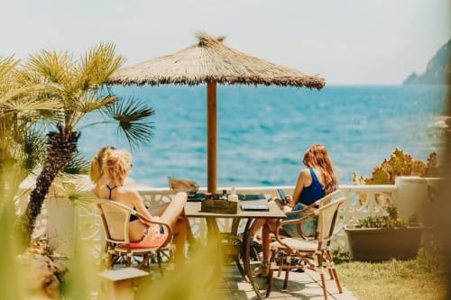 Dos huéspedes trabajando en una mesa frente al mar bajo una sombrilla de paja en Casa Marina Suites, Benissa, Alicante