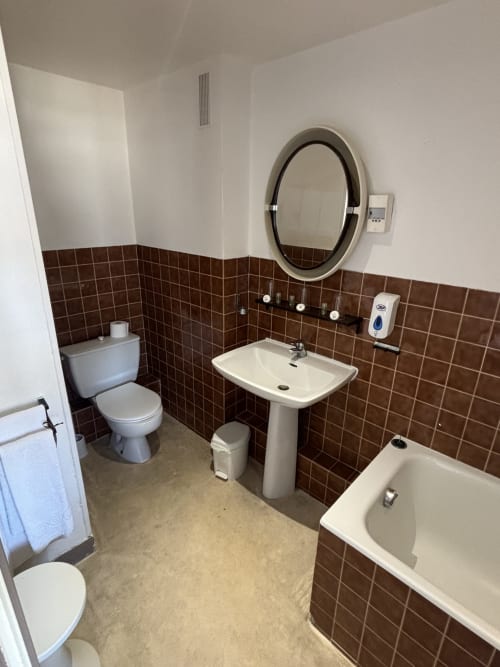 Salle de bain de l'Hôtel bistrot de la poste avec toilettes, lavabo sur colonne, miroir, baignoire et carrelage marron
