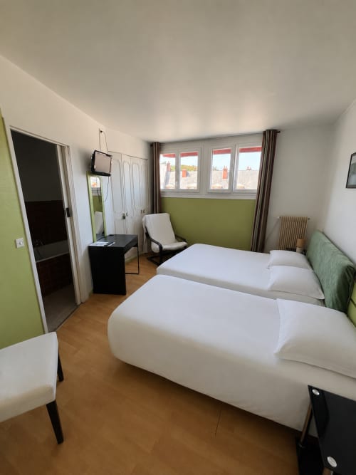 Chambre triple avec deux lits blancs, mur d'accent vert, vue sur la ville et parquet à l'Hôtel bistrot de la poste