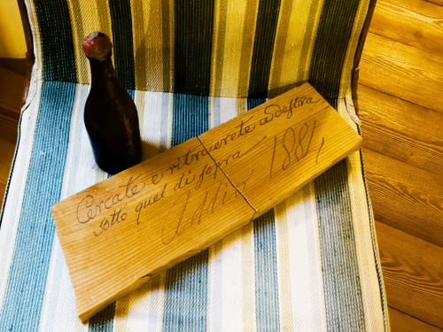 Targa in legno della Roseto del Drago B&B a Ponte in Valtellina con testo inciso, accanto una bottiglia di vino scuro su tessuto rigato