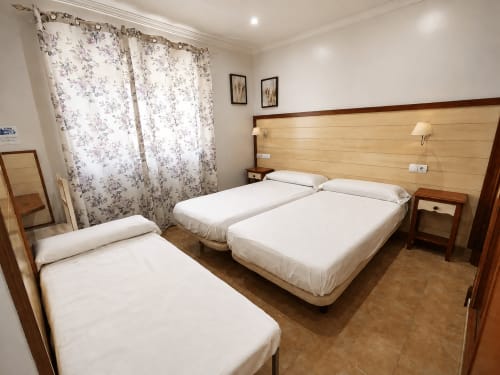 Habitación triple en Hostal La Moyareja de Rota con tres camas, cabecero de madera, cortinas florales y suelo de baldosas