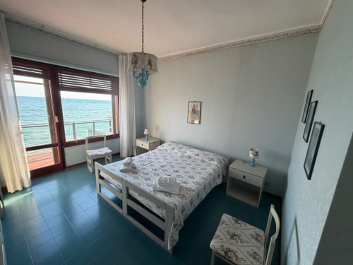 Camera da letto luminosa fronte mare a Villa Mareluce a Santa Marinella con vista sul mare, letto bianco, pavimento in maioliche blu e accesso al balcone