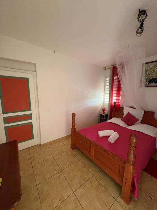 Chambre lumineuse avec lit en bois, literie magenta, serviettes blanches, porte rouge, ventilateur de plafond, sol carrelé à la villa L'Aseta Paradise