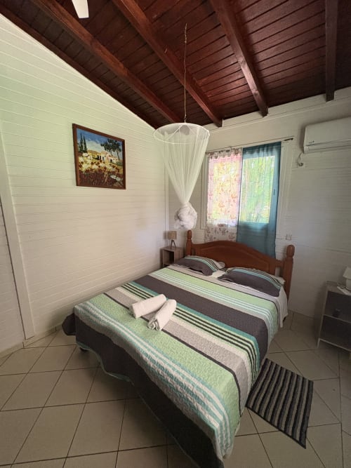 Chambre lumineuse avec literie rayée verte, poutres en bois, fenêtres vue mer et moustiquaire au plafond à la villa L'Aseta Paradise